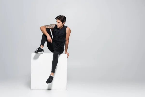 Athletic sportsman in black sportswear sitting on cube on grey background — Stock Photo