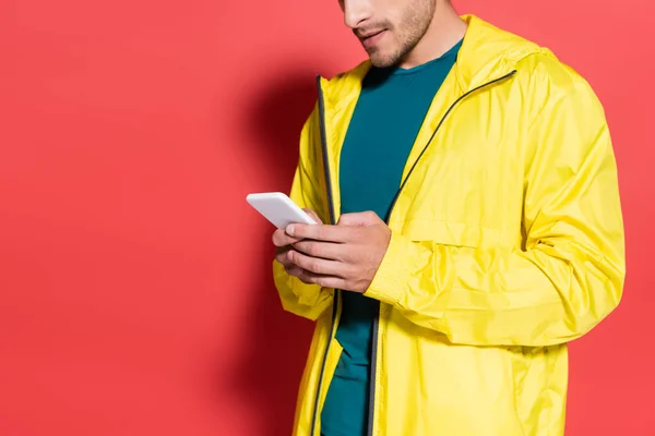 Vista recortada del deportista en chaqueta deportiva amarilla usando teléfono móvil sobre fondo rojo - foto de stock