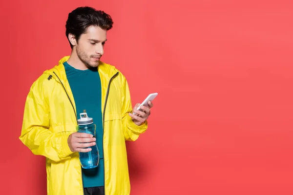 Sportsman holding sports bottle and using smartphone on red background — Stock Photo