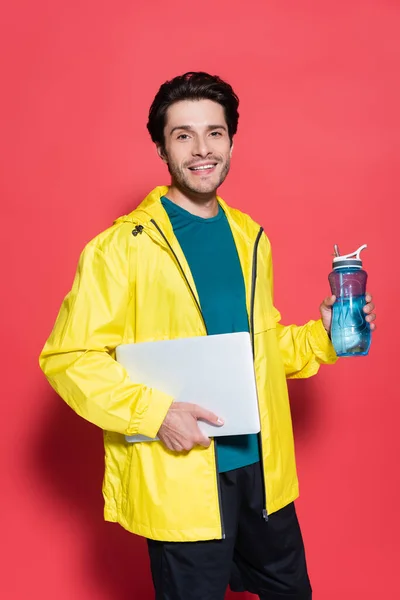 Desportista feliz segurando laptop e garrafa de esportes enquanto olha para a câmera no fundo vermelho — Fotografia de Stock