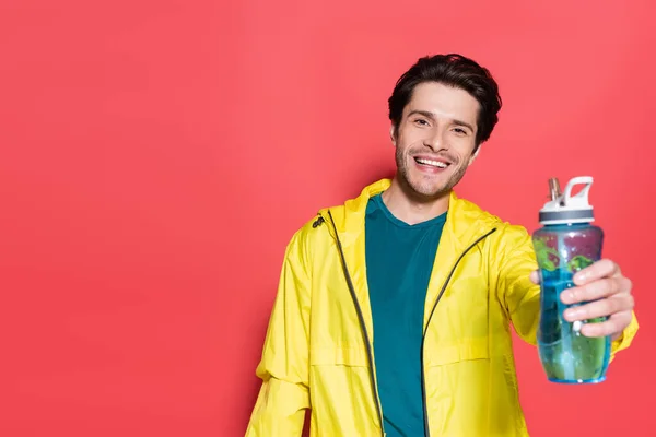 Cheerful sportsman holding blurred sports bottle on red background — Stockfoto