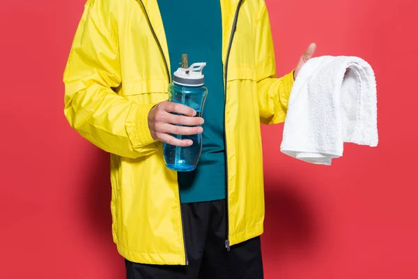 Vista recortada del deportista en chaqueta deportiva sosteniendo agua y toalla sobre fondo rojo - foto de stock