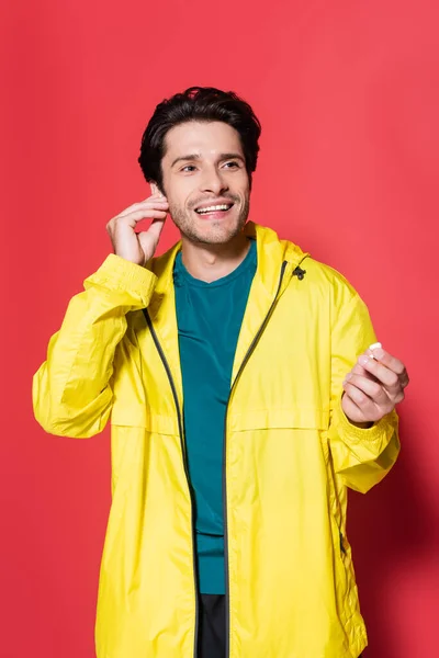 Deportista sonriente en chaqueta deportiva sosteniendo auriculares inalámbricos sobre fondo rojo - foto de stock