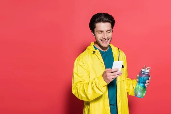 Desportista positivo no fone de ouvido usando telefone celular e segurando garrafa de esportes no fundo vermelho — Fotografia de Stock