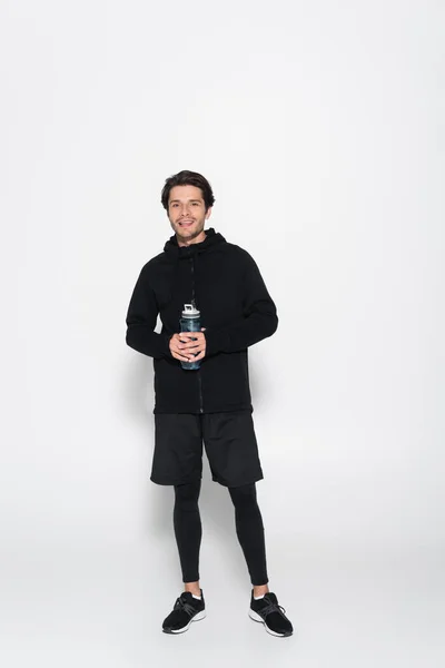 Full length of smiling sportsman holding sports bottle and looking at camera on grey background — Stockfoto