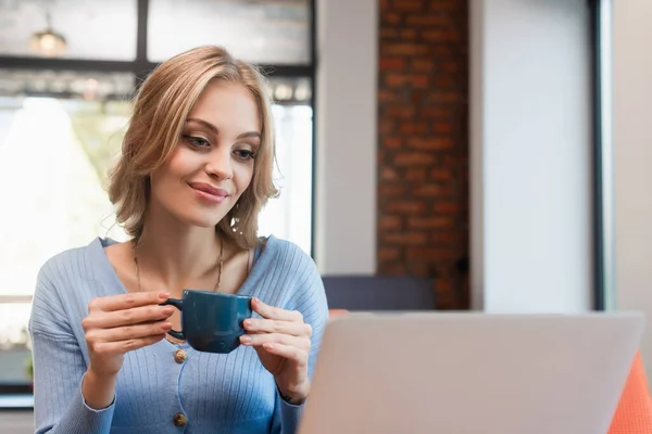 Smiling woman holding cup of coffee while looking at blurred laptop in cafe — Fotografia de Stock