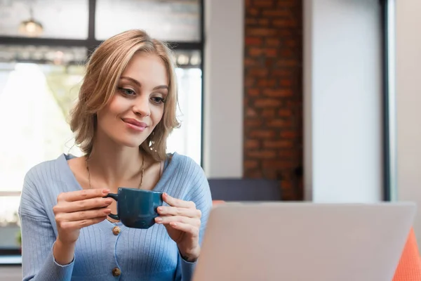 Sonriente mujer sosteniendo la taza de café mientras mira borrosa portátil en la cafetería - foto de stock