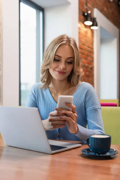 Femme gaie bavarder sur smartphone près d'un ordinateur portable et une tasse de café — Photo de stock