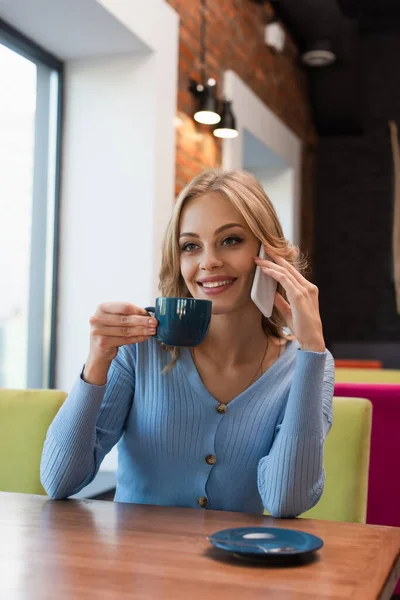 Femme souriante tenant tasse de café tout en parlant sur téléphone mobile dans le café — Photo de stock