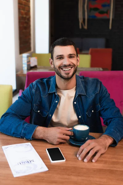 Homem feliz sentado no restaurante com xícara de café perto do menu e smartphone com tela em branco na mesa — Fotografia de Stock