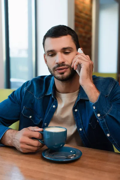 Молодой бородатый мужчина разговаривает по телефону и держит чашку капучино в кафе — стоковое фото