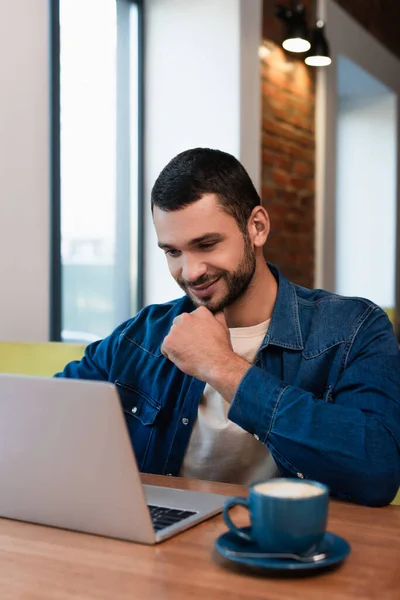 Lächelnder Mann blickt auf Laptop neben verschwommener Kaffeetasse im Café — Stockfoto