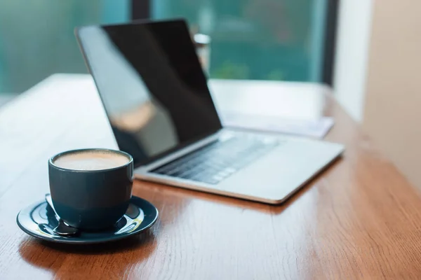 Messa a fuoco selettiva della tazza con cappuccino vicino al computer portatile offuscato con schermo bianco sul tavolo nel caffè — Foto stock