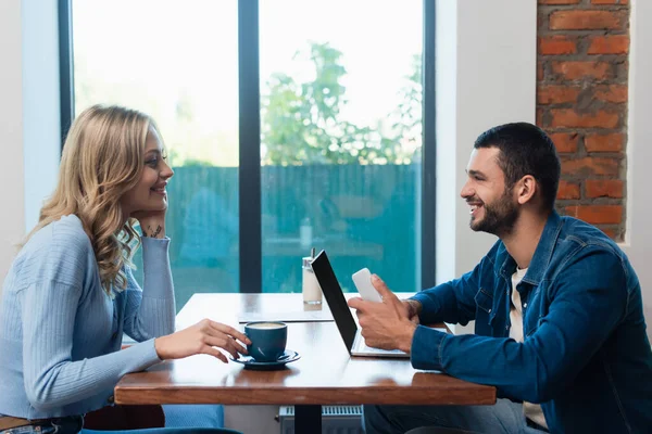 Vista lateral de pareja feliz mirándose el uno al otro cerca de la computadora portátil con pantalla en blanco y taza de capuchino — Stock Photo