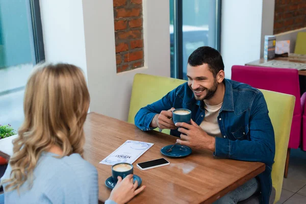 Fröhlicher Mann trinkt Kaffee mit Freundin in der Nähe von Smartphone mit leerem Bildschirm und Speisekarte auf Tisch — Stockfoto