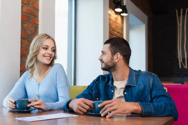 Feliz pareja mirándose mientras se sienta cerca de tazas de café - foto de stock