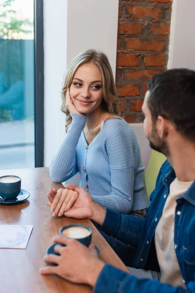 Mujer alegre cogida de la mano con el hombre borroso cerca de tazas de café en la mesa - foto de stock