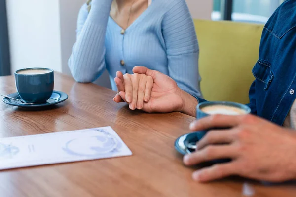 Vista parcial de pareja cogida de la mano cerca de tazas de café y menú borroso - foto de stock