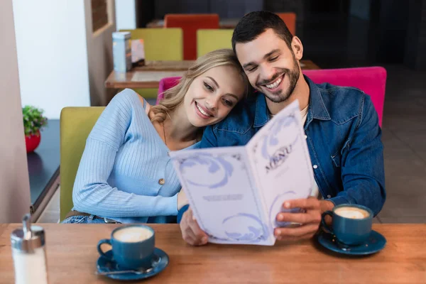 Alegre pareja elegir la comida de menú cerca de tazas de capuchino - foto de stock