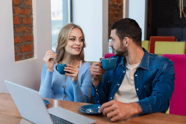 Happy couple with coffee cups looking at each other near blurred laptop in restaurant — Stock Photo