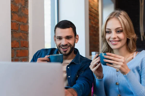 Веселая пара с кофейными чашками, смотрящая на размытый ноутбук, проводя время в кафе — стоковое фото