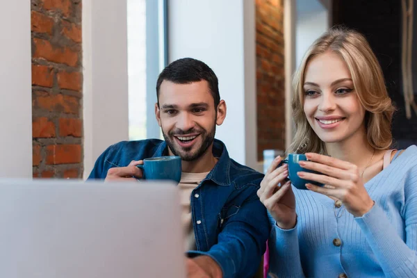 Cheerful couple with coffee cups looking at blurred laptop while spending time in cafe — Fotografia de Stock