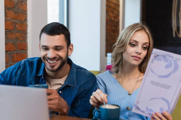 Homem sorrindo olhando para laptop borrado perto de mulher escolhendo refeição de menu no restaurante — Fotografia de Stock