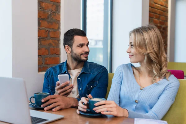 Jeune homme avec smartphone regardant petite amie souriante près des tasses avec cappuccino et ordinateur portable flou — Photo de stock