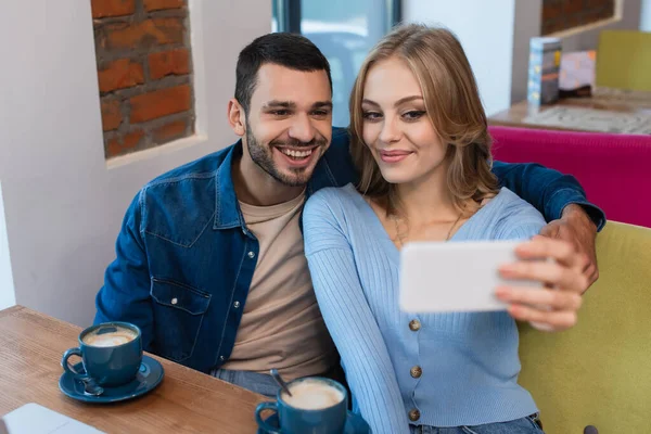 Donna bionda che prende selfie con fidanzato felice vicino a tazze di caffè, primo piano sfocato — Foto stock