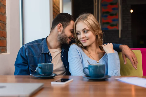 Happy man embracing girlfriend near blurred coffee cups, smartphone and laptop in restaurant — Fotografia de Stock