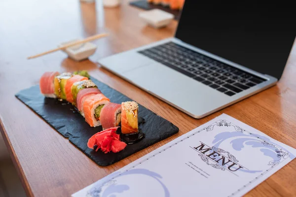 Deliciosos rollos de sushi, menú y computadora portátil borrosa con pantalla en blanco en la mesa en el bar de sushi - foto de stock