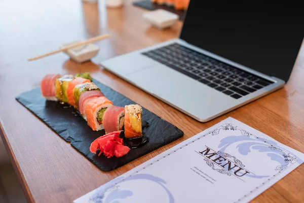 Delicious sushi rolls, menu and blurred laptop with blank screen on table in sushi bar — Stock Photo