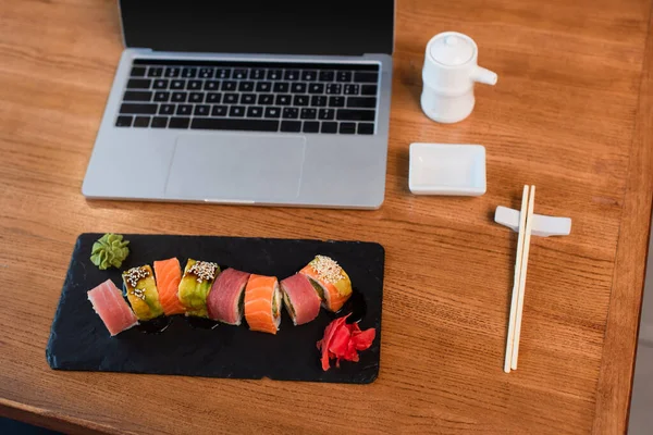 Vista de alto ângulo da placa com rolos de sushi, pauzinhos, panela de molho de soja e laptop com tela em branco na mesa de madeira — Fotografia de Stock