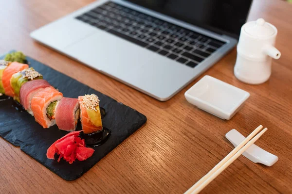 Sabrosos rollos de sushi, palillos, olla de salsa de soja y un tazón cerca de la computadora portátil borrosa en la mesa en el bar de sushi - foto de stock