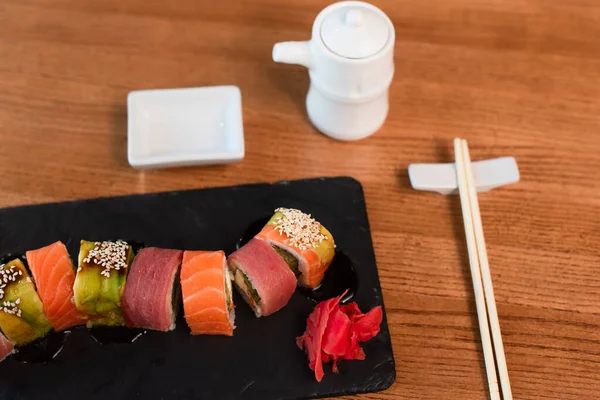 Rolos de sushi frescos perto de pauzinhos, panela de molho de soja turva e tigela na mesa de madeira — Fotografia de Stock
