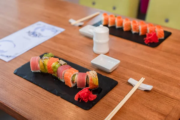 Pratos pretos com rolos de sushi tradicionais perto de pauzinhos, conjunto de molho de soja e menu borrado na mesa no bar de sushi — Fotografia de Stock