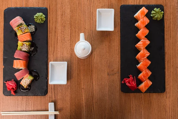 Top view of black plates with delicious sushi rolls near chopsticks, soy sauce pot and bowl on wooden table — Stock Photo