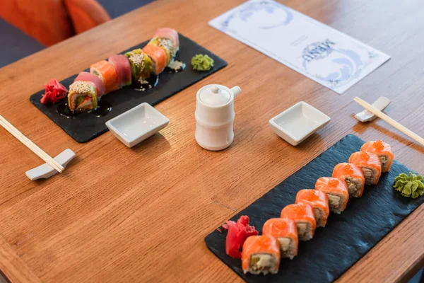Pratos pretos com deliciosos rolos de sushi com wasabi e gengibre perto do pote de molho de soja e folheto de menu na mesa de madeira — Fotografia de Stock