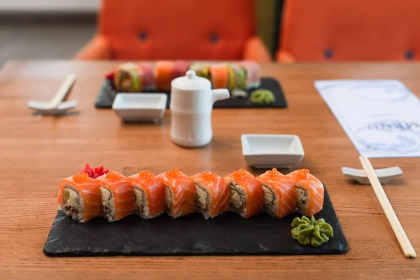Assiettes noires avec de savoureux rouleaux de sushi près des baguettes, casserole de soja et menu flou sur table en bois — Photo de stock
