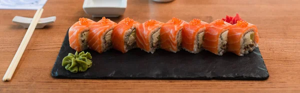 Plaque noire avec ensemble de rouleaux de sushi près de wasabi et baguettes sur table en bois, bannière — Photo de stock