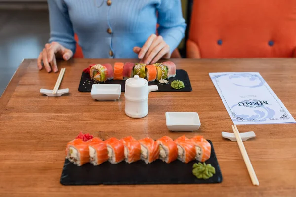Vue recadrée de femme floue près du menu, assiettes avec rouleaux de sushi, baguettes, casserole de soja et bols — Photo de stock