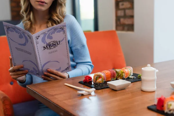 Vista ritagliata della donna offuscata menu di lettura vicino gustosi rotoli di sushi sul tavolo — Foto stock