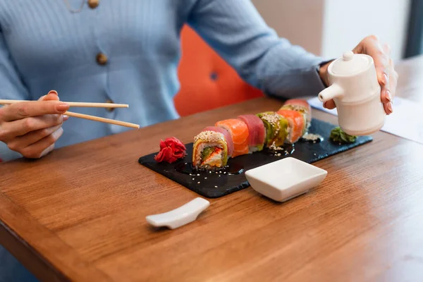 Partial view of blurred woman holding chopsticks and soy sauce pot near delicious sushi rolls — Stockfoto