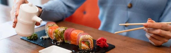 Vue recadrée d'une femme floue tenant des baguettes et un pot de sauce soja près des rouleaux de sushi, bannière — Photo de stock