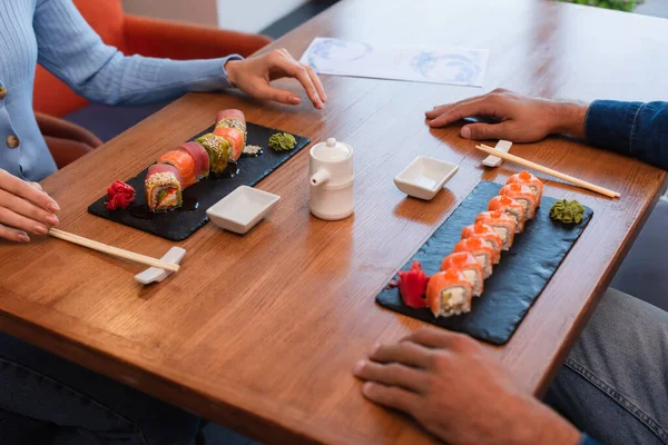 Cropped view of couple near table with delicious sushi, chopsticks, soy sauce pot and bowls — Stockfoto