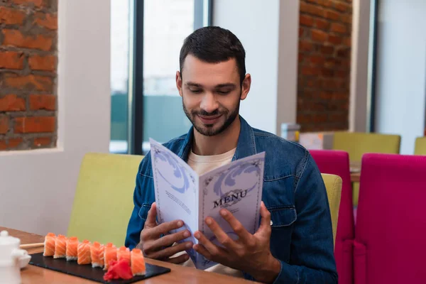 Un homme souriant lisant le menu près de la plaque avec des rouleaux de sushi au restaurant — Photo de stock