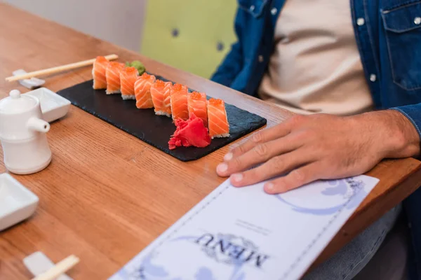 Vista recortada del hombre cerca del plato de rollos de sushi y menú borroso en la mesa - foto de stock