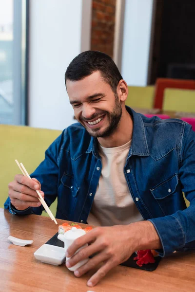 Homme joyeux riant avec les yeux fermés tout en tenant des baguettes près des rouleaux de sushi — Photo de stock