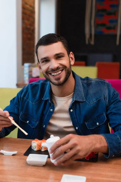 Joven sonriendo a la cámara mientras sostiene palillos y salsa de soja cerca de rollos de sushi - foto de stock