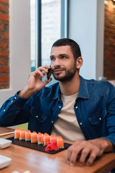 Улыбающийся мужчина разговаривает по мобильному телефону рядом с тарелкой с суши в ресторане — стоковое фото